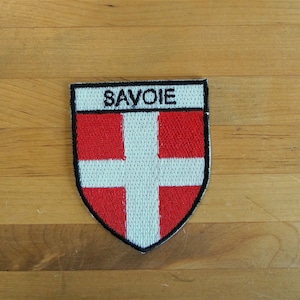 Peut inclure: Écusson brodé représentant les armoiries de la Savoie. L'écusson en forme de bouclier présente une croix blanche sur fond rouge, avec le mot "SAVOIE" en lettres blanches en haut. L'écusson est bordé de noir.