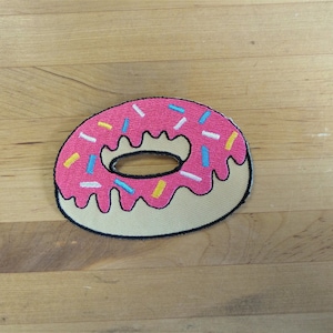 May include: A pink frosted donut with colorful sprinkles. The donut is embroidered on a beige fabric.