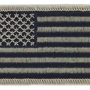 Könnte beinhalten: Ein kleiner, gestickter amerikanischer Flaggen-Patch mit einem braunen Rand. Die Sterne und Streifen sind in einem Tarnmuster mit braunen und schwarzen Farben gehalten.