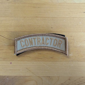 Puede incluir: Un parche de tela beige con la palabra "CONTRACTOR" bordada en hilo dorado.