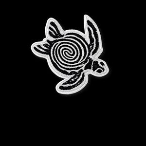 Op de afbeelding: Een zwart-wit geborduurde patch in de vorm van een zeeschildpad. De schild van de schildpad heeft een spiraalpatroon en de randen zijn wit. De patch staat op een zwarte achtergrond.