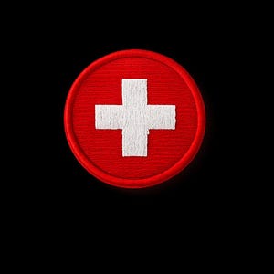 Op de afbeelding: Een rode, ronde patch met een wit kruis in het midden. De patch heeft een gestikte rand en staat tegen een zwarte achtergrond. Het ontwerp is eenvoudig en gedurfd.