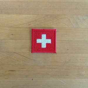May include: A red square patch with a white embroidered cross in the center. The patch is a Swiss flag.