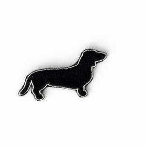 Peut inclure: Un écusson noir en forme de chien teckel avec un contour blanc cousu. L'écusson représente un chien teckel, une race connue pour son long corps et ses courtes pattes. L'écusson est sur fond blanc.