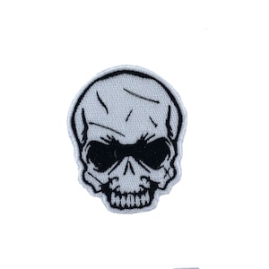 Pode incluir: Etiqueta branca bordada com um design de caveira preta. A caveira tem um design detalhado com a testa franzida e uma expressão ameaçadora.
