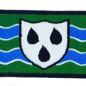 Peut inclure: Écusson brodé avec un bouclier blanc et trois formes de goutte noires, sur un fond ondulé vert et bleu. L'écusson a une bordure noire, créant une forme rectangulaire. Pour vêtements ou accessoires.
