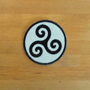 May include: A white circular patch with a black triple spiral design. The patch is embroidered with a white background and black thread.