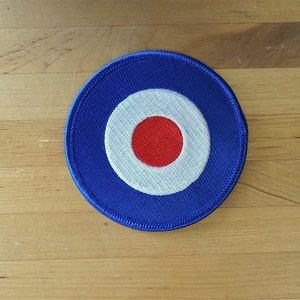 Peut inclure: Un patch de cible bleu, blanc et rouge. La cible est un cercle avec un anneau blanc autour d'un centre rouge.