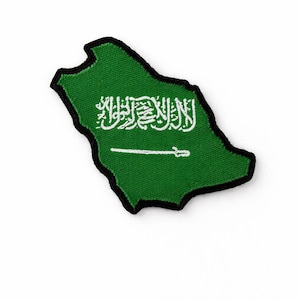 Puede incluir: Un parche verde con la forma de Arabia Saudí, con un borde negro. El parche presenta escritura árabe blanca y una espada, que representa el emblema nacional. El fondo es de color verde liso.