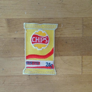 May include: An embroidered patch of a bag of crisps. The bag is yellow and red and says "CHIPS" with a red and white stripe across the bottom.