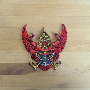 May include: A red, yellow, and blue embroidered patch depicting the Garuda, a mythical bird-like creature, with outstretched wings. The Garuda is a symbol of Thailand.