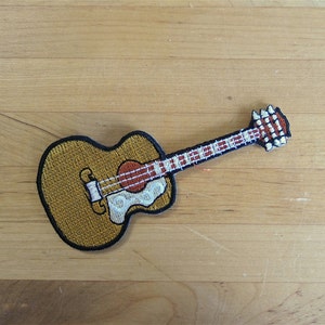 May include: A brown and gold embroidered patch of an acoustic guitar with a white pickguard.