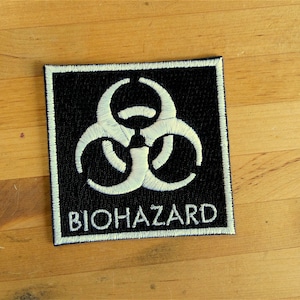 Könnte beinhalten: Ein schwarzer quadratischer Patch mit einem weißen Biohazard-Symbol und dem Wort "BIOHAZARD" in weiß.