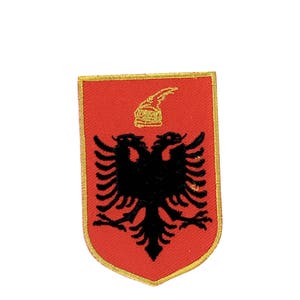 May include: A red embroidered patch with a gold outline, featuring a black double-headed eagle with a gold crown on top.