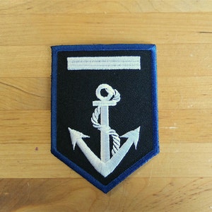 Può includere: Un'ancora bianca ricamata su una toppa nera con bordo blu. L'ancora ha un dettaglio di corda ed è circondata da una striscia bianca.