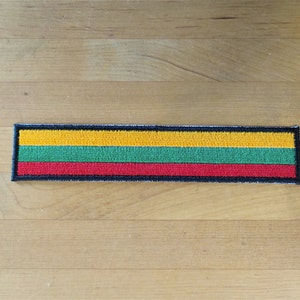 May include: A yellow, green, and red embroidered patch with a black border. The patch is shaped like a rectangle and is on a wooden surface.