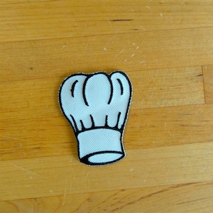 Può includere: Toppa ricamata a forma di cappello da chef bianco con contorni neri. La toppa è su una superficie di legno.