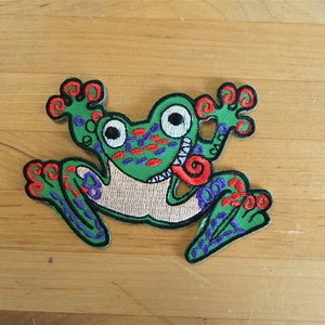 Puede incluir: Parche de rana bordado en verde, rojo y azul con la lengua enroscada. La rana tiene un diseño caprichoso y de dibujos animados.