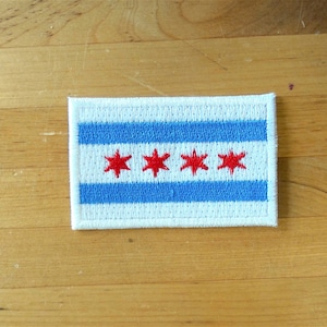 Puede incluir: Un parche blanco con un diseño a rayas azul y blanco y cuatro estrellas rojas en el centro. Las estrellas están dispuestas en una fila.