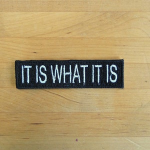 Puede incluir: Parche bordado en blanco y negro con el texto "IT IS WHAT IT IS" sobre una superficie de madera.
