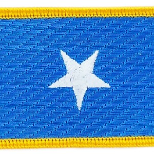 May include: A blue fabric patch with a white five-pointed star in the center. The patch is surrounded by a yellow border.