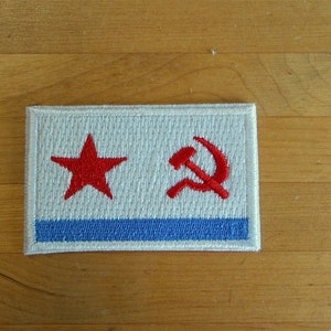 May include: A white patch with a red star and a red hammer and sickle embroidered on it. The patch has a blue stripe across the bottom.