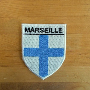 Può includere: Toppa ricamata bianca e blu con una forma di scudo bianco e una croce blu. Il testo "MARSEILLE" è ricamato in nero nella parte superiore dello scudo.