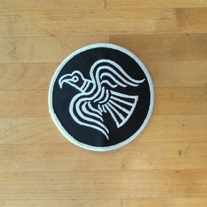 May include: A black and white embroidered patch of a stylized raven with its wings spread. The patch is circular and has a white border.