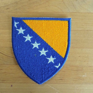 Peut inclure: Un écusson brodé bleu et jaune en forme de bouclier avec huit étoiles blanches. Les étoiles sont disposées en demi-cercle sur le côté bleu de l'écusson.
