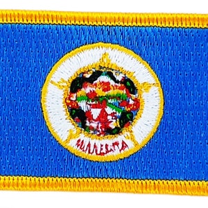 Pode incluir: Um remendo azul com uma borda amarela e um círculo branco no centro. O círculo contém um selo com um design vermelho, branco e verde e a palavra "MINNESOTA" em um arco amarelo.