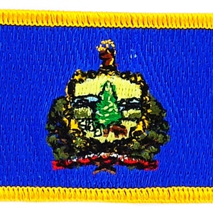 Può includere: Toppa ricamata della bandiera dello stato del Vermont. La bandiera è blu con un pino verde e un sigillo dorato al centro. Il sigillo è circondato da una ghirlanda di foglie verdi e un nastro rosso. La toppa ha un bordo giallo.