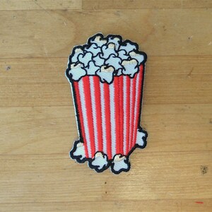May include: A red and white striped popcorn bucket with white popcorn inside. The bucket is embroidered with white stitching.