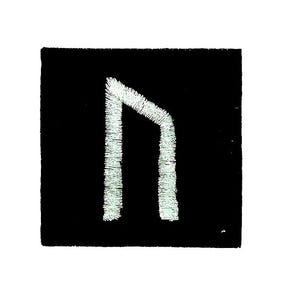 Peut inclure: Un écusson carré noir avec un symbole de rune brodé en blanc. La rune est un motif angulaire stylisé, possiblement une lettre ou un symbole d'un alphabet ancien. La broderie blanche crée un contraste texturé sur le fond noir.
