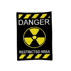Op de afbeelding: Zwart rechthoekig embleem met gele en witte geborduurde tekst en afbeeldingen. Het embleem toont het woord "DANGER" boven een geel stralingssymbool. Onder het symbool staat de tekst "RESTRICTED AREA". Gele diagonale strepen omlijsten de boven- en onderkant.