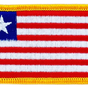 Può includere: Una toppa ricamata della bandiera liberiana. La bandiera è rossa, bianca e blu con una stella bianca nel cantone blu. La toppa è circondata da un bordo giallo.