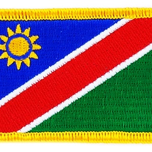 May include: A rectangular embroidered patch featuring the flag of Namibia. The flag is blue with a yellow sun in the upper left corner, a red diagonal stripe with a white border, and a green lower right corner.