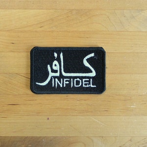 May include: Black rectangular patch with white text in Arabic script and the English word "INFIDEL". The patch is likely made of fabric and is set against a light brown wooden surface.