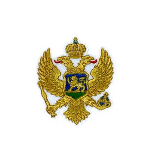 Puede incluir: Un parche bordado en oro que representa un águila bicéfala que sostiene un cetro y un orbe. El águila tiene un escudo en el centro, que es azul con un león y un fondo verde.