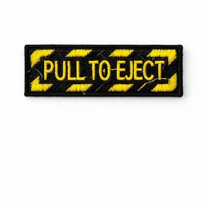 Op de afbeelding: Zwart rechthoekig embleem met gele zigzagranden en de geborduurde tekst "PULL TO EJECT" in geel. De patch is een waarschuwings- of instructielabel, waarschijnlijk voor een apparaat of kleding.