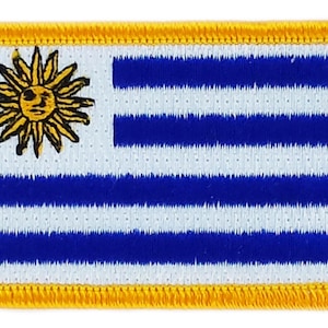Pode incluir: Um remendo bordado da bandeira uruguaia. A bandeira é branca com nove listras horizontais azuis e um sol dourado com um rosto humano no canto superior esquerdo. O remendo é rodeado por uma borda amarela.