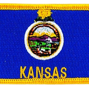 May include: A blue embroidered patch with a yellow border. The patch features the state seal of Kansas, which depicts a sun rising over a landscape with a river and a sunflower. The text "KANSAS" is embroidered in yellow below the seal.