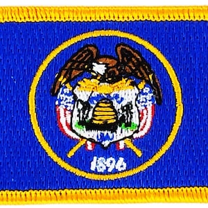 Puede incluir: Un parche bordado azul con un borde amarillo. El parche presenta el sello del estado de Utah, que incluye un águila calva, una colmena y la fecha 1896.