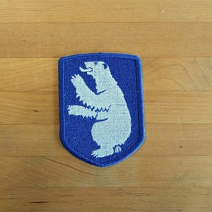 Op de afbeelding: Een blauwe viltpatch met een witte ijsbeer die op zijn achterpoten staat. De beer kijkt naar links en heeft een witte omtrek.