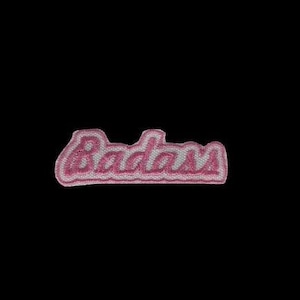 May include: A pink and white embroidered patch with the word "Badass" in a cursive font. The patch has a white outline and a pink fill, set against a black background.