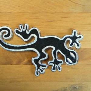 Pode incluir: Etiqueta de gecko bordada em preto com costuras brancas. O gecko está enroscado com a cauda em espiral.