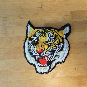 Op de afbeelding: Een geborduurde tijgerkop-patch in geel en zwart op een witte achtergrond. De tijger heeft een woeste uitdrukking met scherpe tanden en een rode tong.