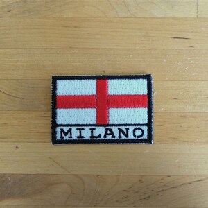 Puede incluir: Un parche bordado blanco y rojo con la palabra "MILANO" en letras negras.