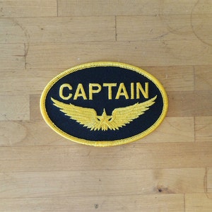 May include: A black oval patch with yellow stitching. The patch has a yellow embroidered winged design with a star in the center and the word "CAPTAIN" in yellow lettering.