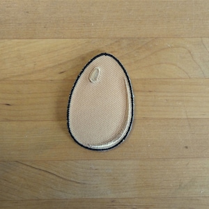 May include: A beige fabric egg-shaped patch with a black outline.