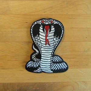Könnte beinhalten: Weiß gestickter Kobra-Patch mit roter Zunge und schwarzem Umriss. Der Patch befindet sich auf einer Holzoberfläche.